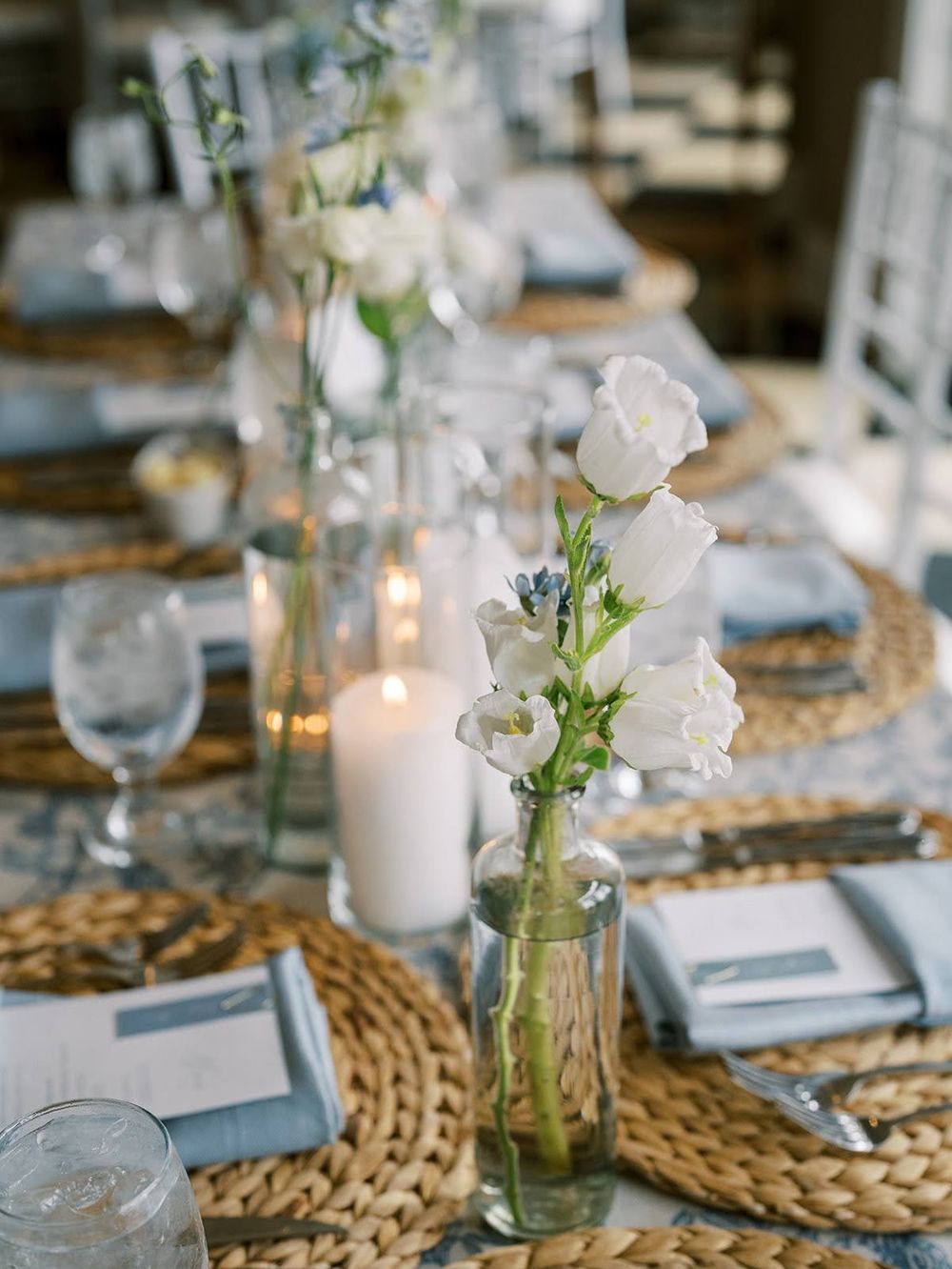 Elegant wedding table with white flowers in glass vases, pillar candles, woven placemats, and soft blue place settings.