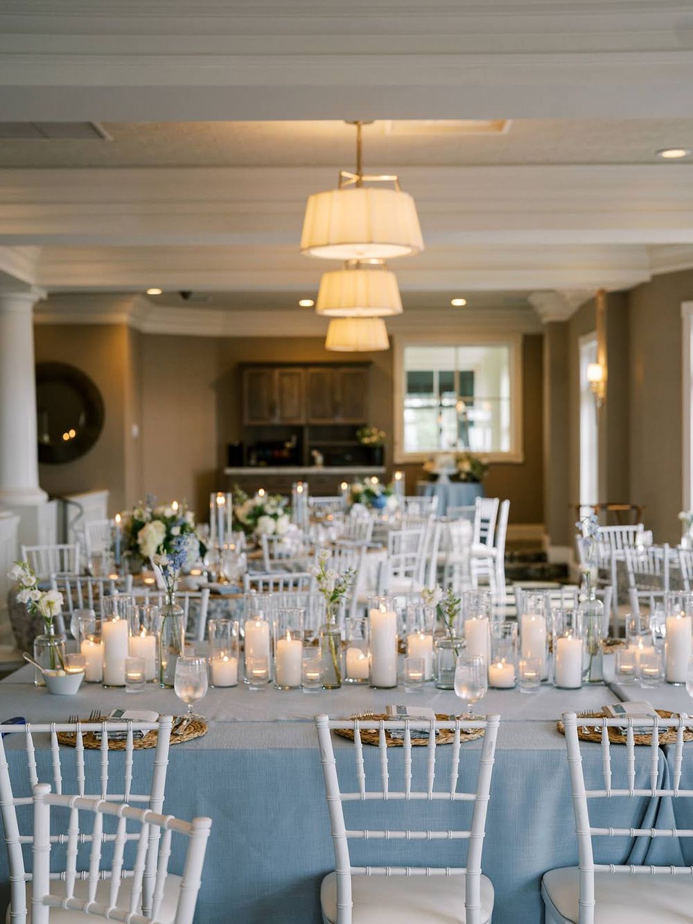 Elegant wedding reception with dusty blue linens, white chiavari chairs, and candle centerpieces in a bright venue.