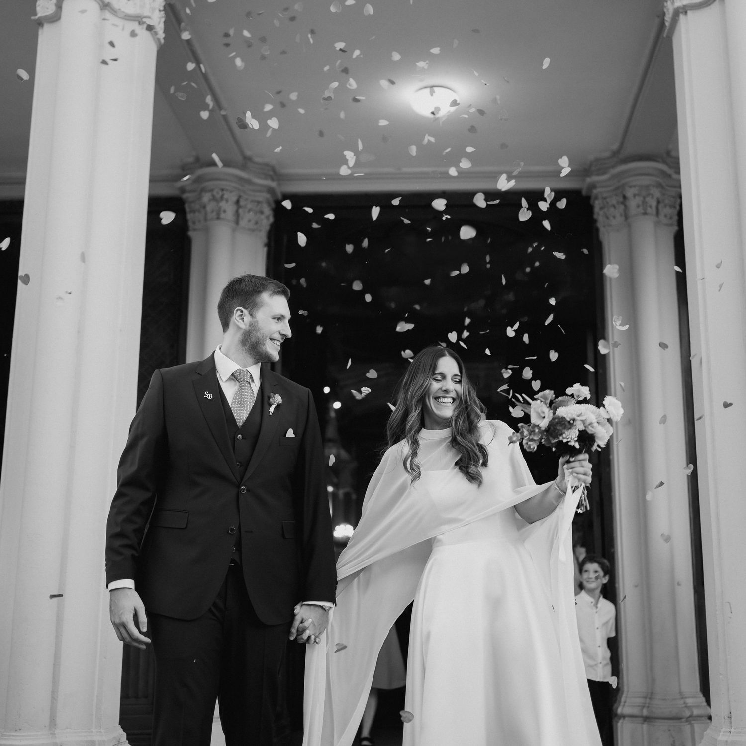 Black and white photo of a bride and groom holding hands, laughing as confetti falls outside a grand columned building.