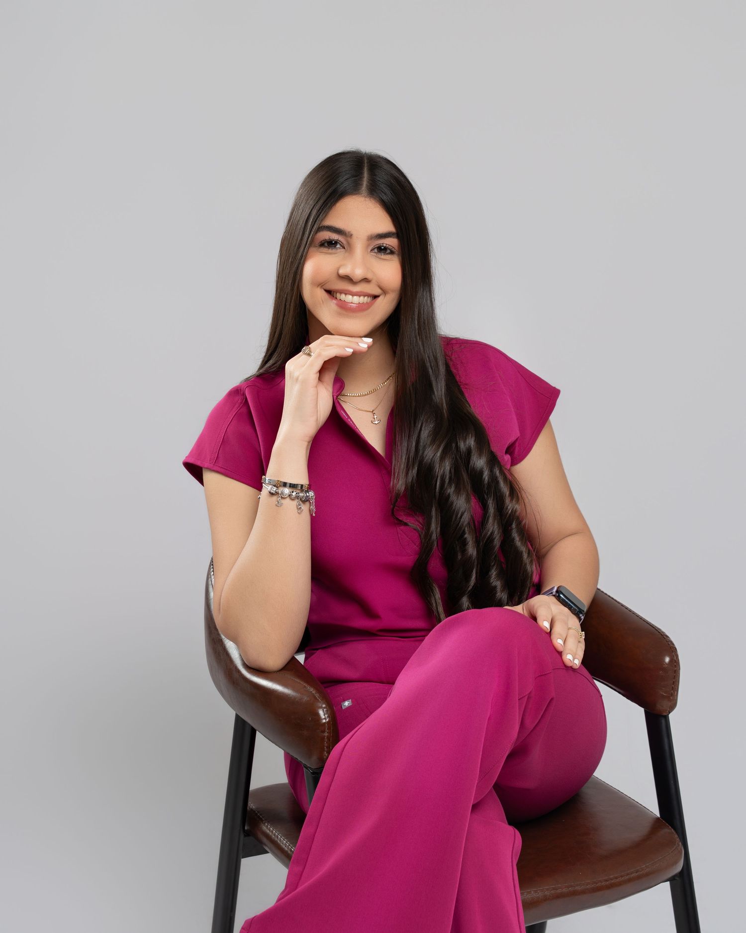 Woman in beige medical scrub jumpsuit poses with stethoscope beside black stool on gray background.