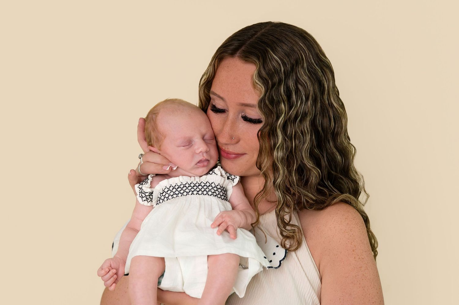 A mother with curly hair tenderly holds her sleeping newborn baby dressed in white during a studio portrait session.