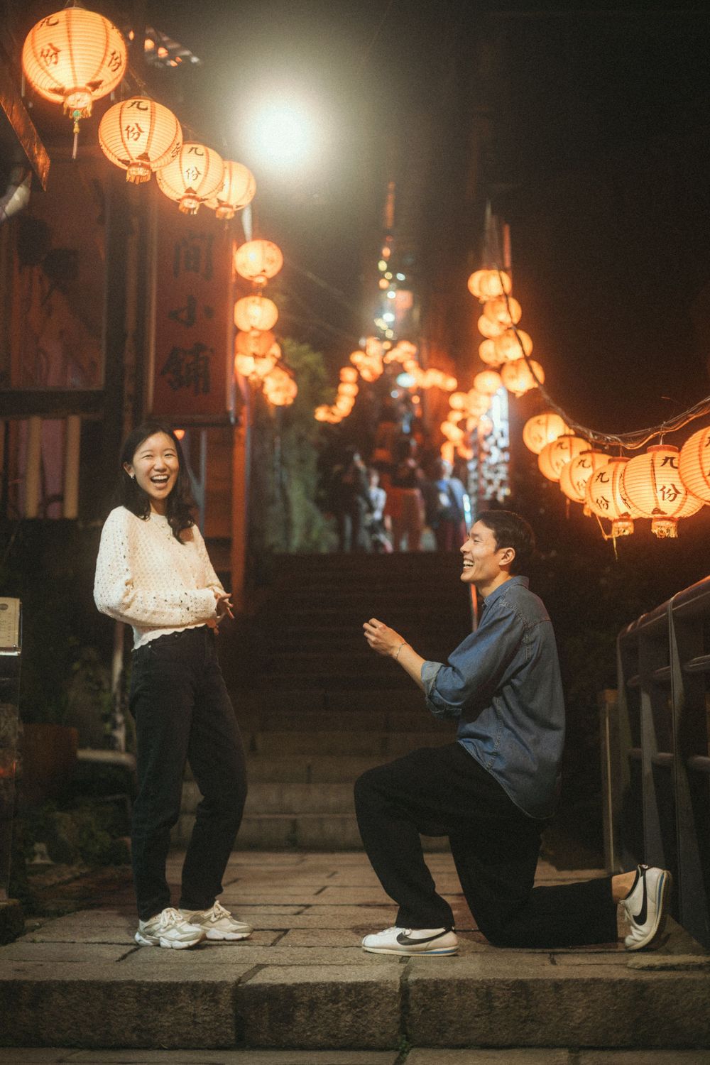 Man proposing to woman on one knee at night, surrounded by glowing red lanterns on stone steps.