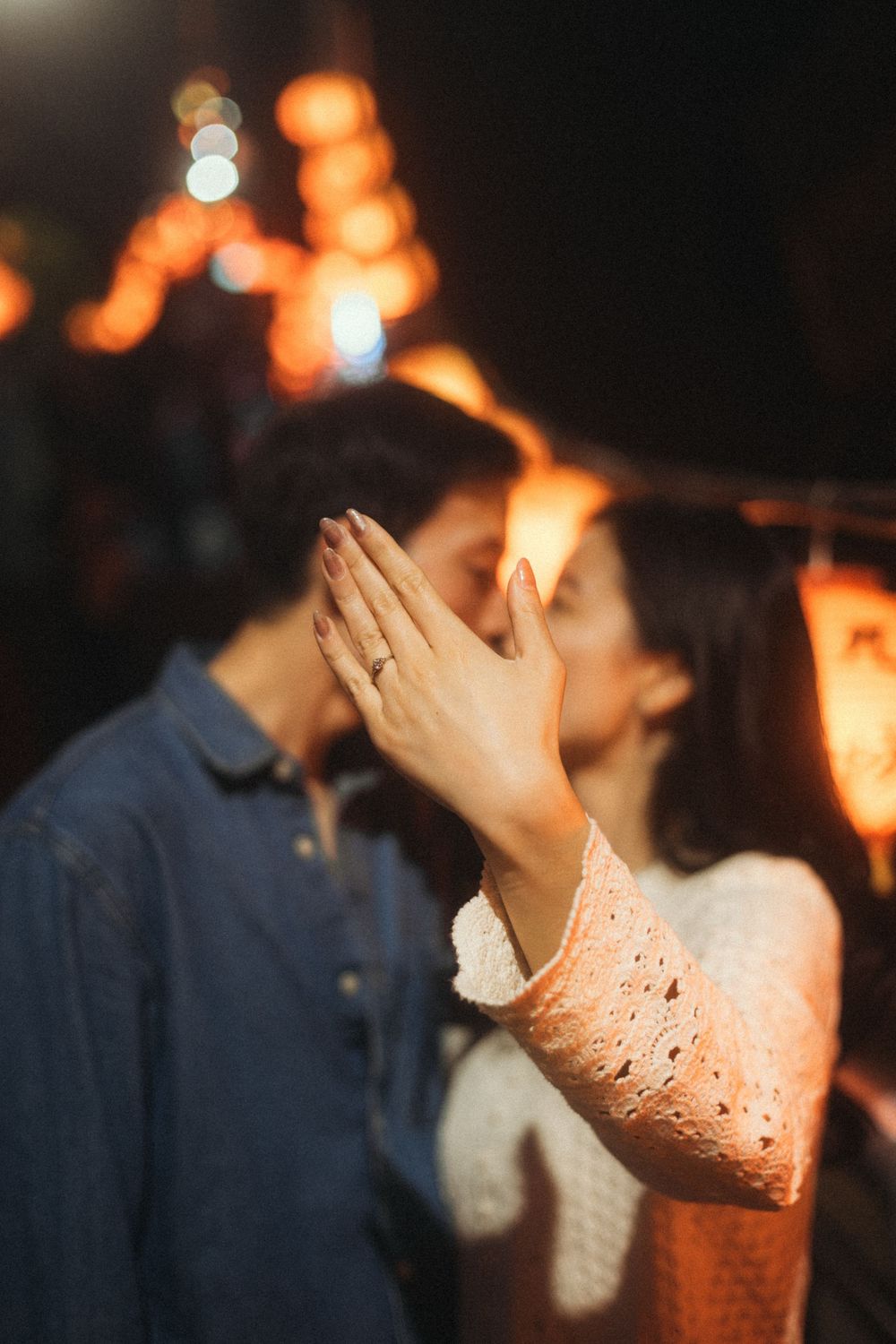 Couple sharing a kiss at night market, woman displaying engagement ring on raised hand with bokeh lights.