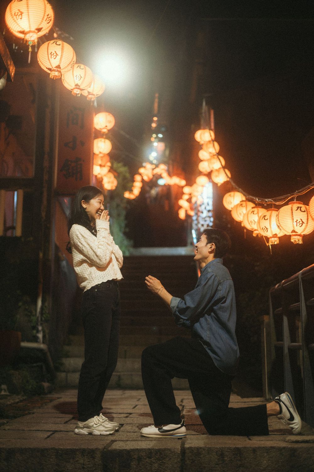 Man proposes on one knee to woman at night under glowing Chinese lanterns on stone steps.