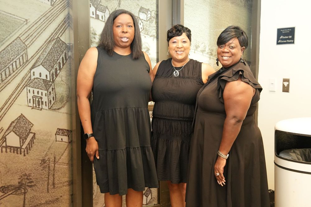 Three women in matching black dresses pose together smiling in front of a historical mural display.