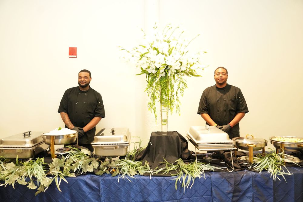 Two catering staff in black chef uniforms stand behind a buffet table with chafing dishes and floral centerpiece.