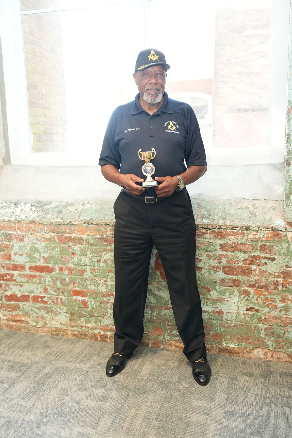 Elderly man in Masonic cap and navy polo holds trophy, standing before rustic brick wall outdoors.