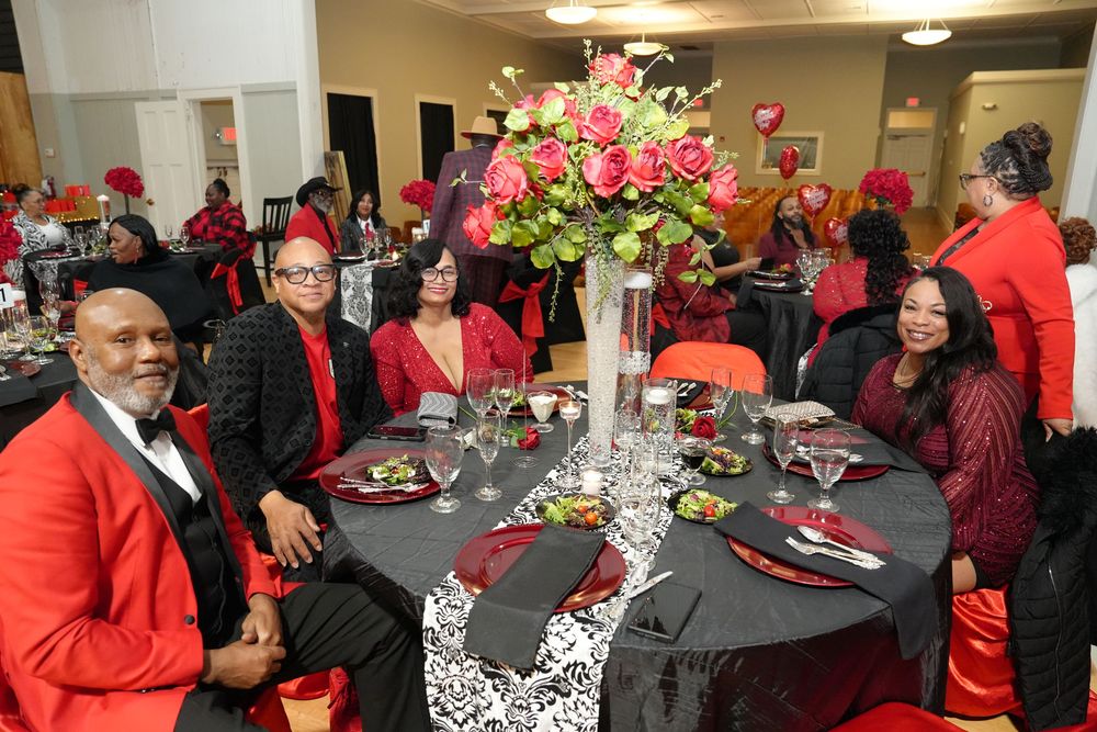 Elegantly dressed guests in red and black attire enjoy a formal gala dinner with floral centerpieces.