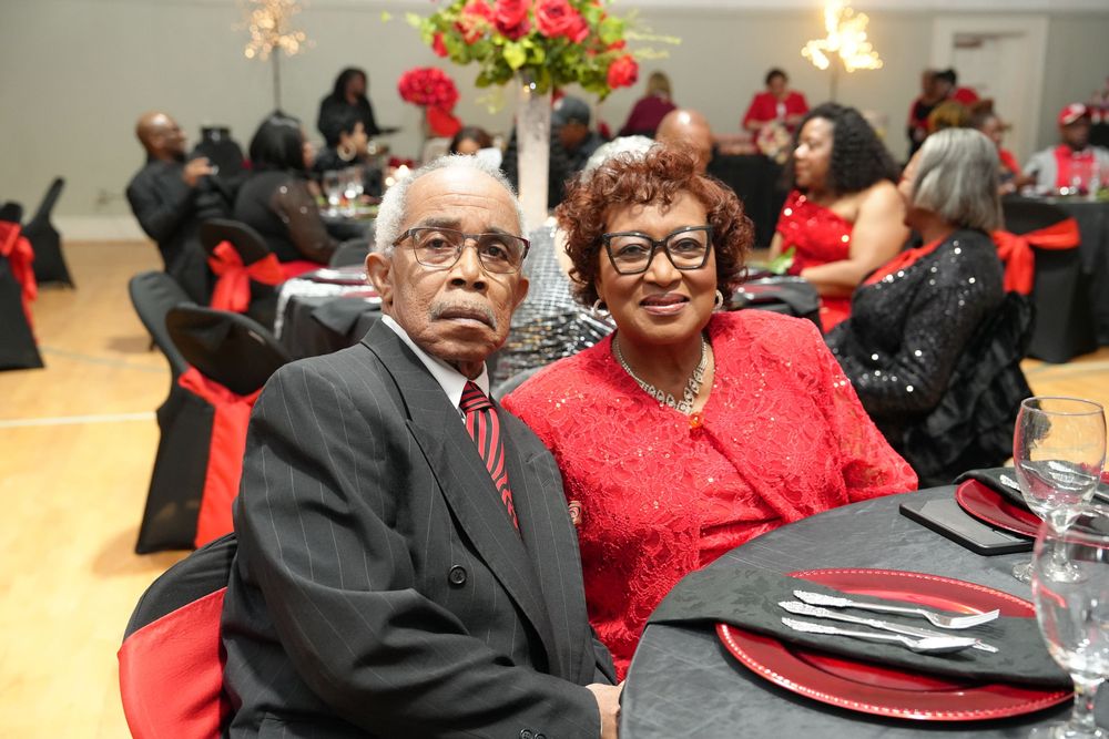 Elderly couple dressed in formal attire seated at a banquet table decorated in red and black at a gala event.