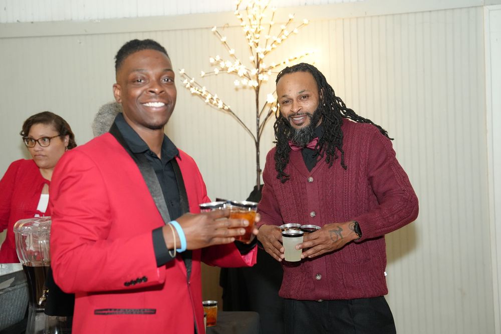 Two men smiling and toasting drinks at a social event, one wearing a red blazer and the other in a maroon shirt.