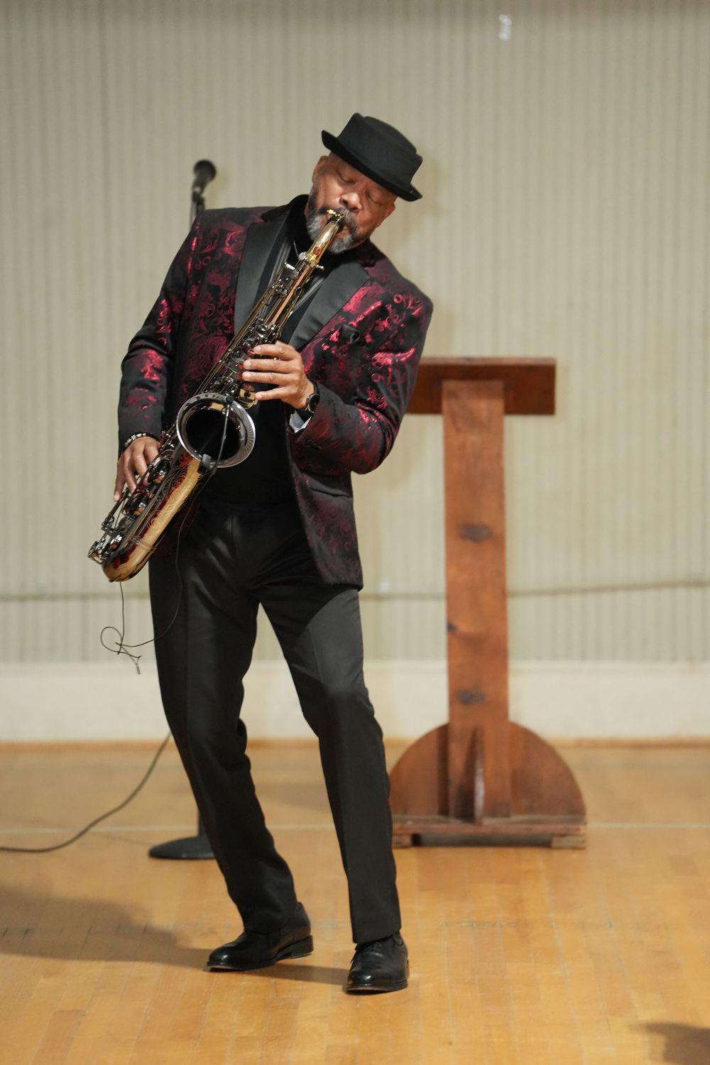 A jazz musician in a red floral blazer and black hat plays saxophone on a wooden stage floor.