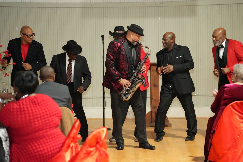 Live jazz band performing with saxophonist in red blazer and cowboy hat at a festive indoor event.