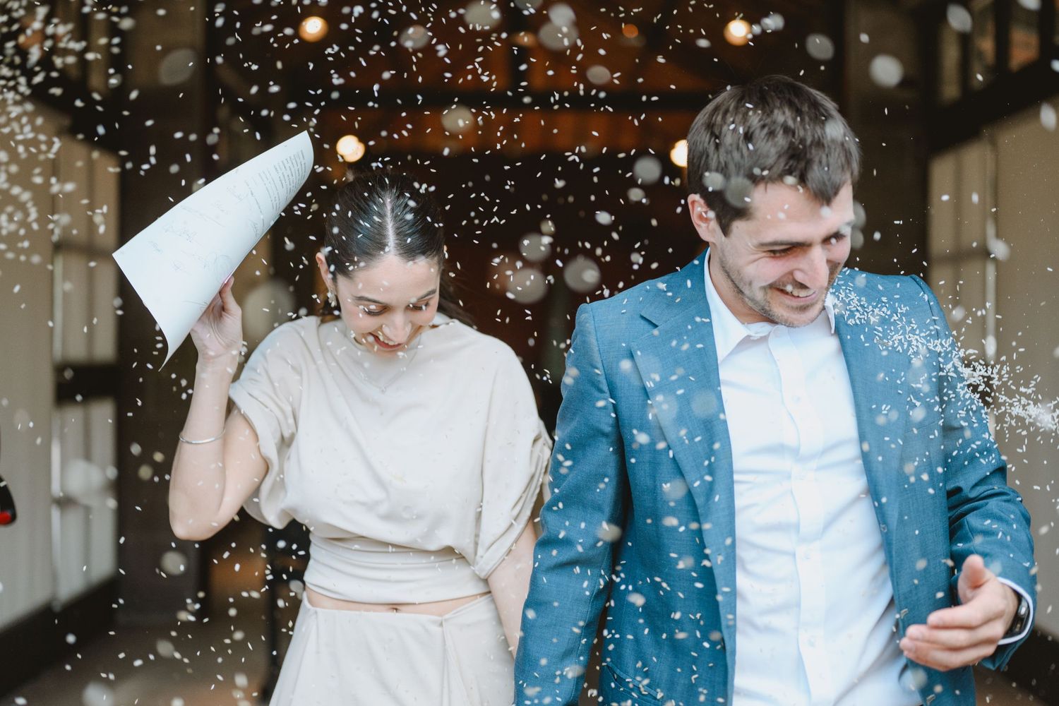 Joyful couple celebrating with confetti shower, woman holding paper cone, man in blue suit smiling outdoors.