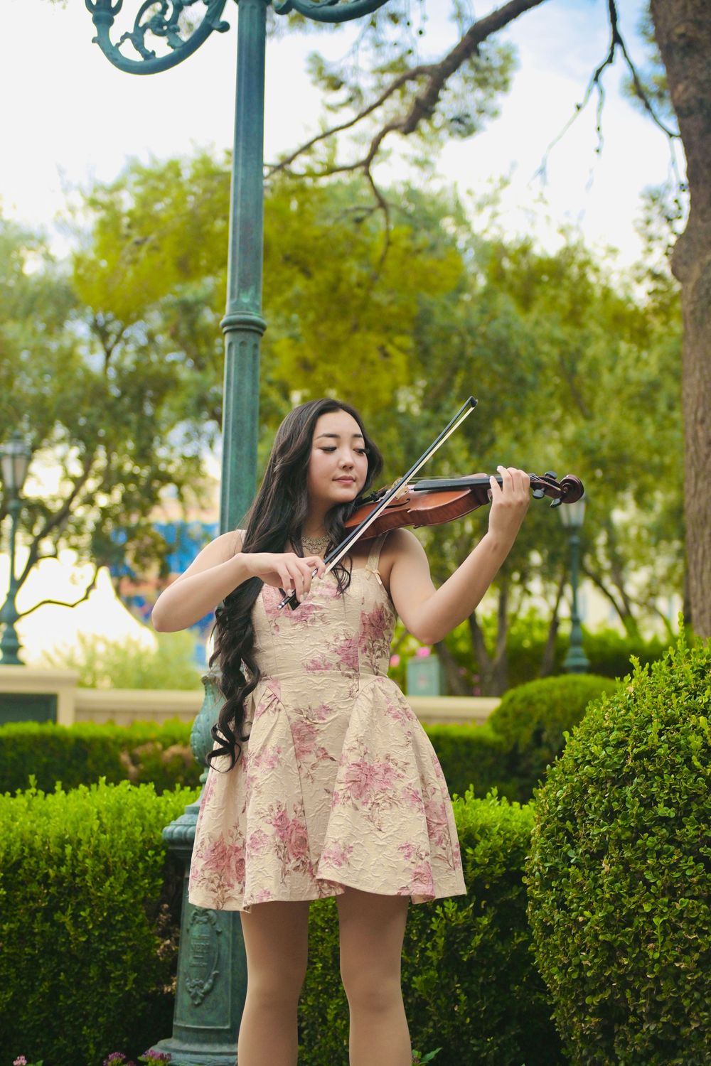 Young Asian woman in floral dress playing violin in a lush green park near a decorative lamp post.