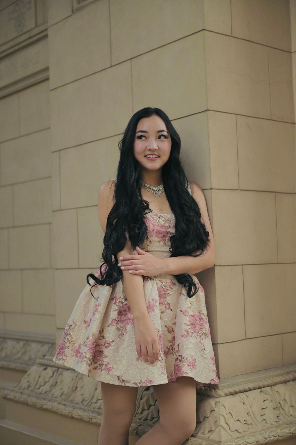 Young woman in floral strapless dress with long black hair posing against a stone wall.