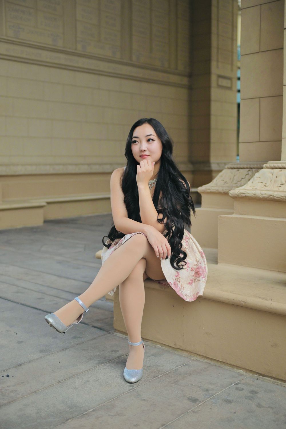 Young Asian woman in floral skirt and silver heels sitting thoughtfully on stone steps of elegant building.