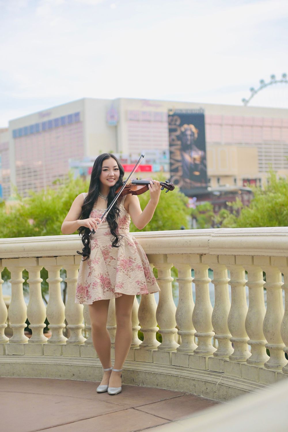 Young woman in floral dress playing violin on ornate balcony with Las Vegas Strip skyline in background.