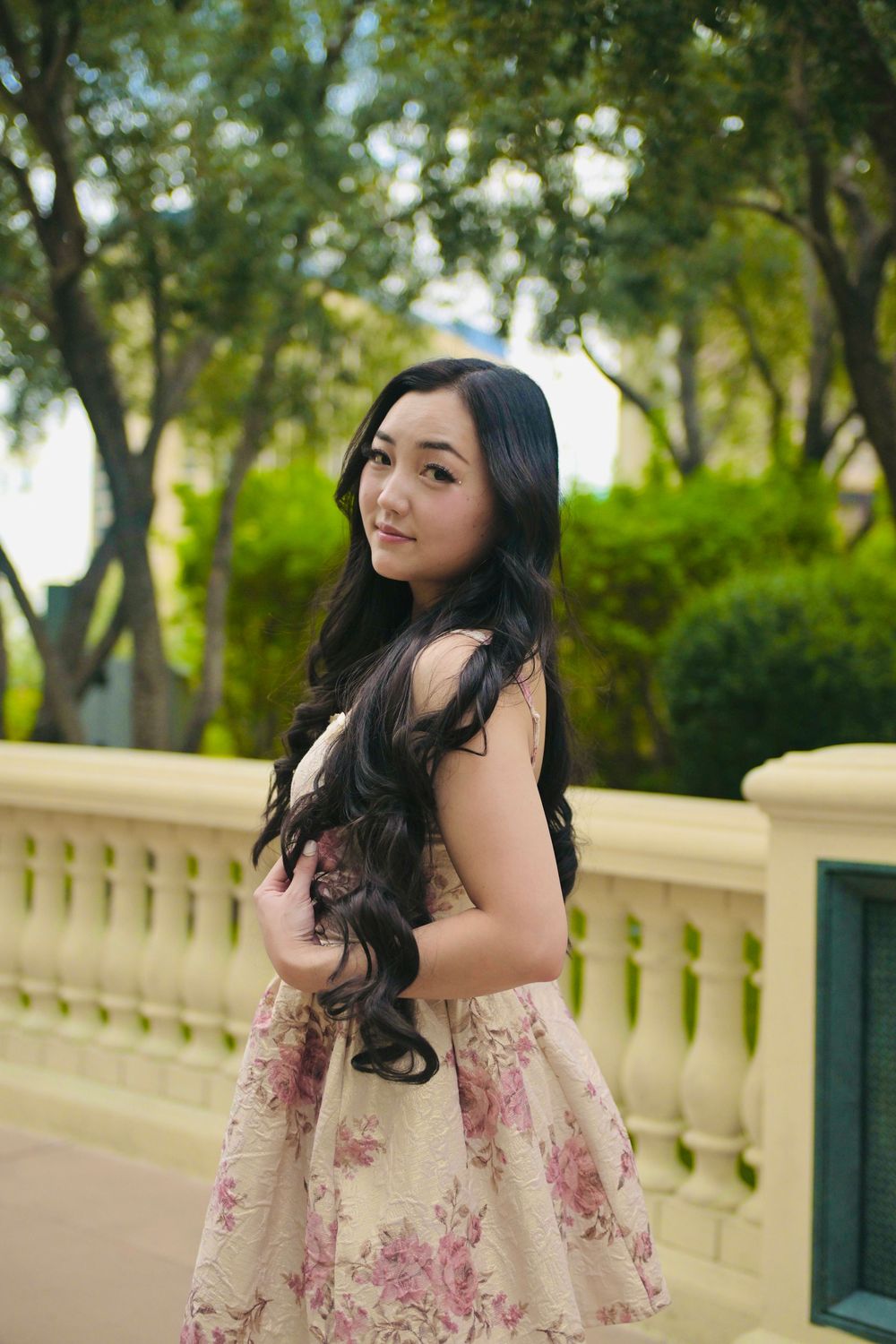 Young Asian woman in floral dress poses gracefully by stone balustrade in lush green park setting.
