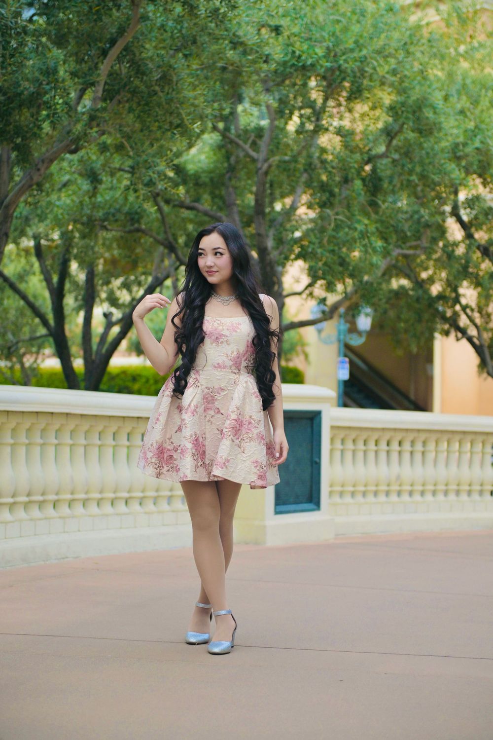 Young woman in floral pink dress with long black hair posing outdoors near white railing with green trees.
