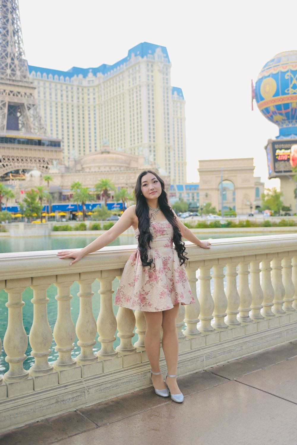 Young Asian woman in floral dress posing by balustrade with Paris Las Vegas hotel and Eiffel Tower replica behind her.