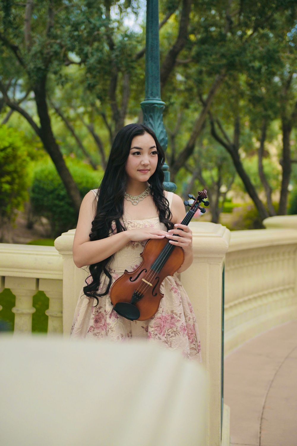 Young woman in floral dress holding violin outdoors by decorative railing with green trees in background.