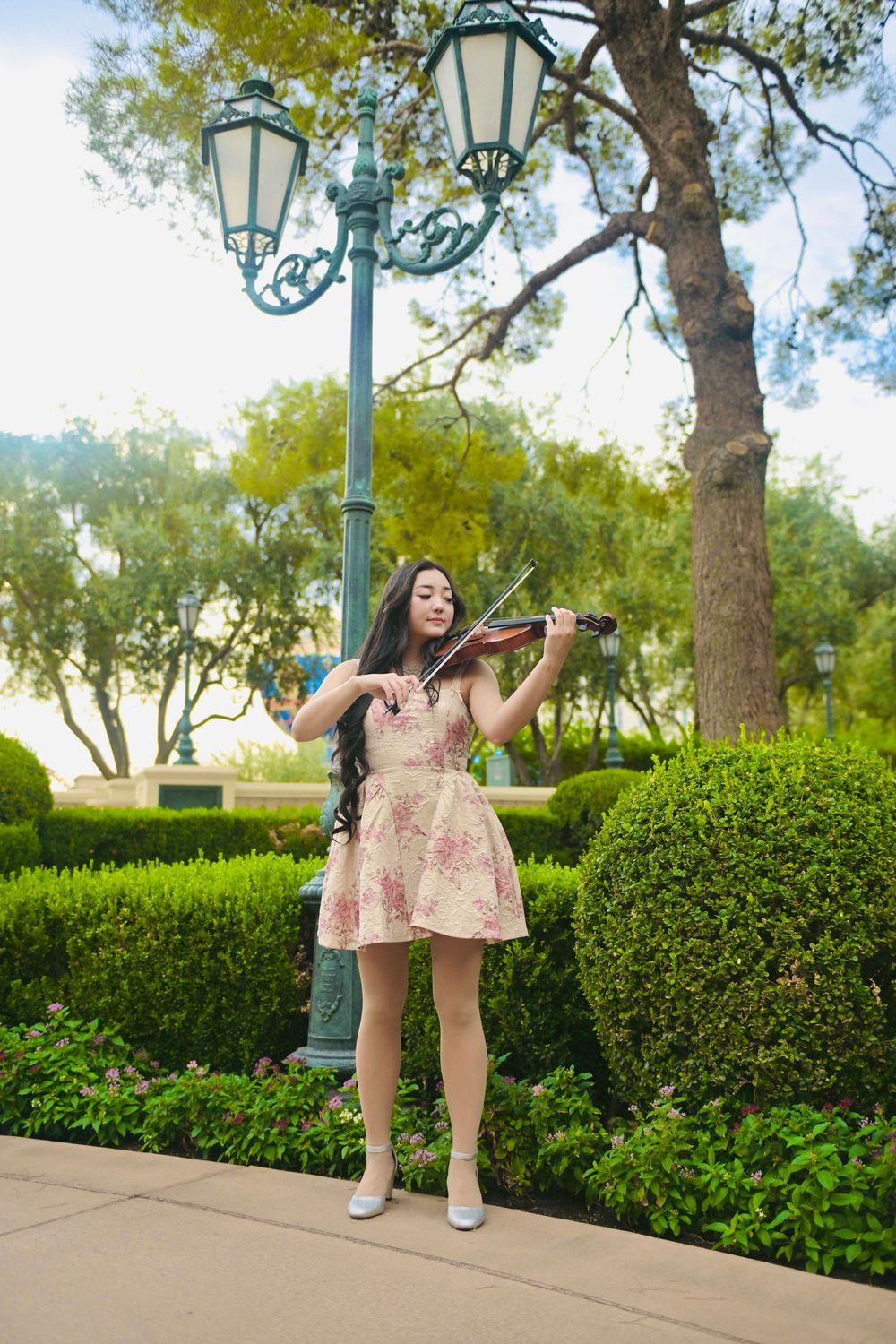 Young woman in floral dress playing violin outdoors in a lush green park near a decorative lamppost.