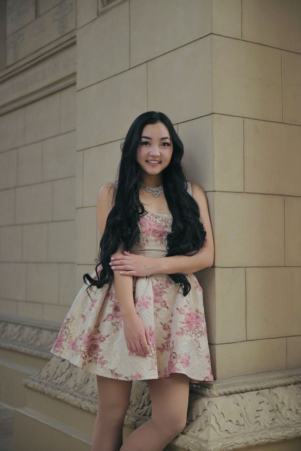 Young Asian woman in floral dress leaning against stone wall, smiling.