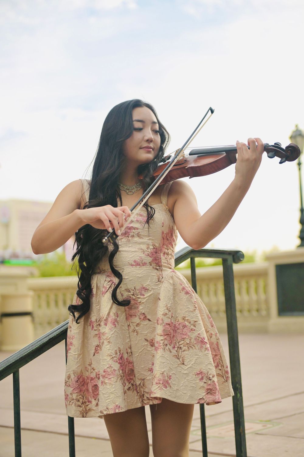 Young woman in floral dress playing violin outdoors on steps with railing.