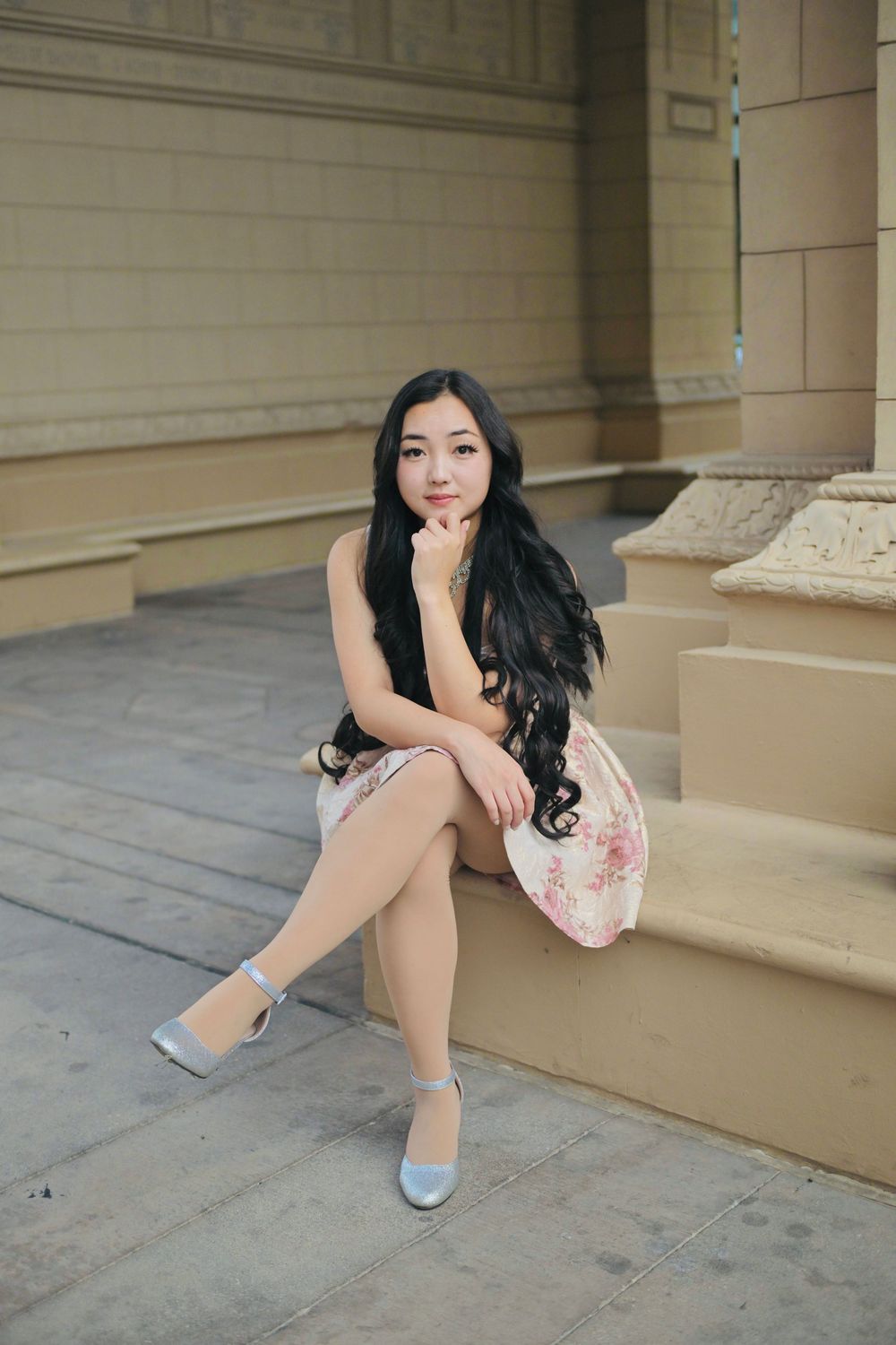 Young Asian woman in floral dress and heels sitting gracefully on stone steps of a classical building.