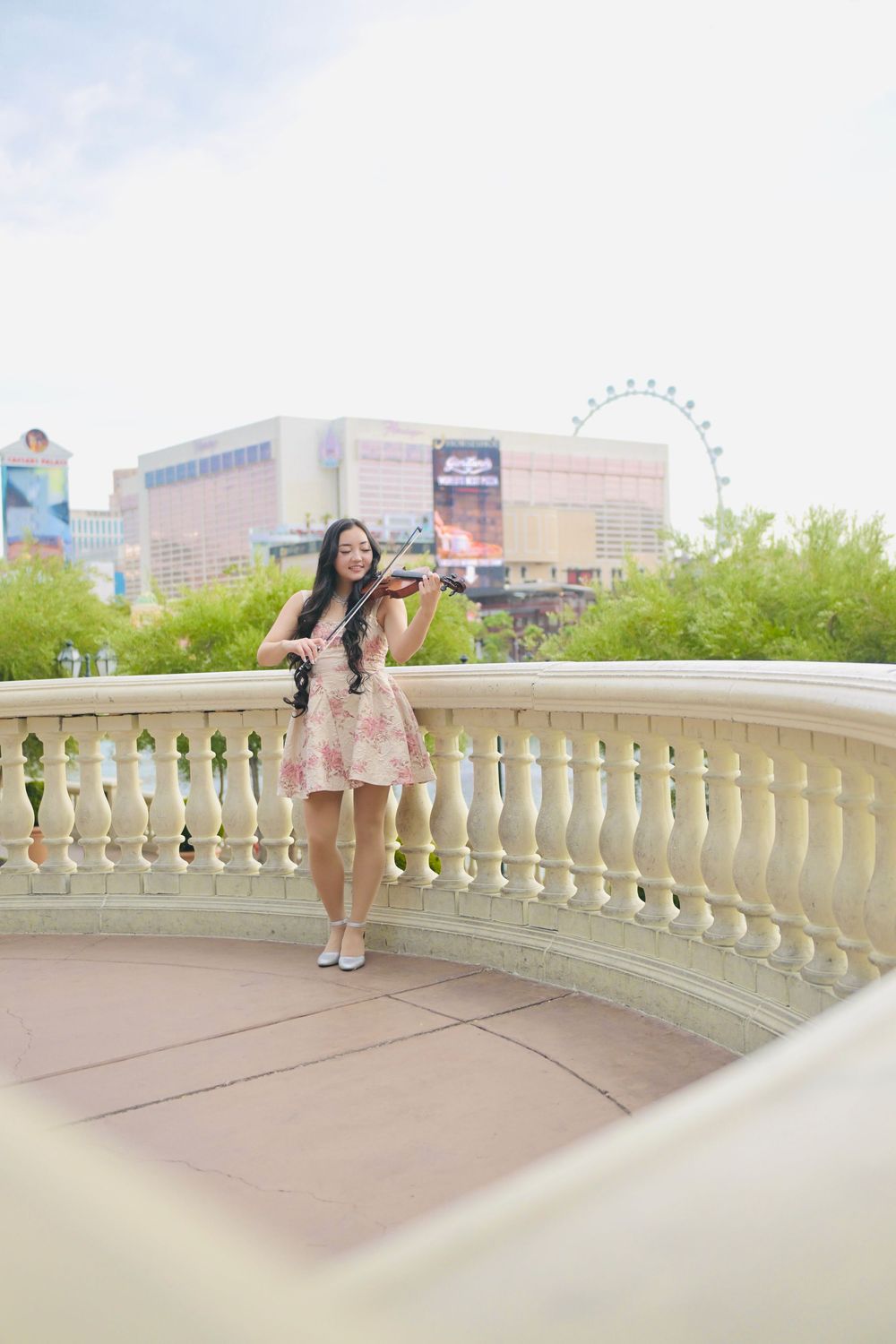 Young woman in floral dress playing violin on elegant balcony with Las Vegas Strip skyline backdrop.