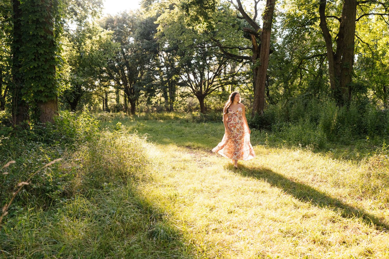 Woman in floral maxi dress walking on sunlit forest path surrounded by lush green trees and tall grass.
