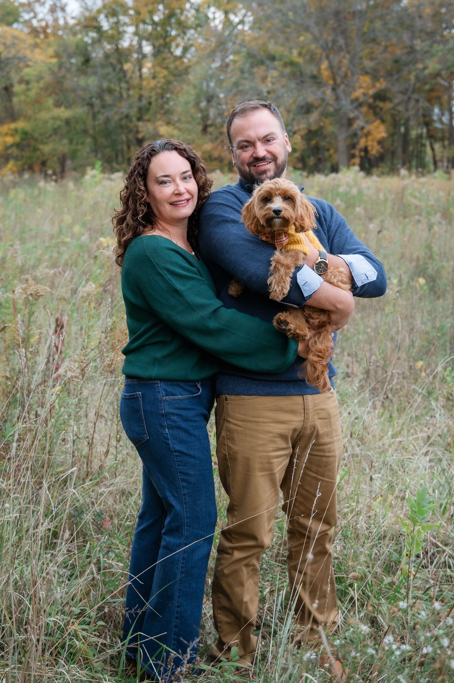 A smiling couple stands in a field holding a fluffy golden doodle dog, surrounded by autumn foliage.