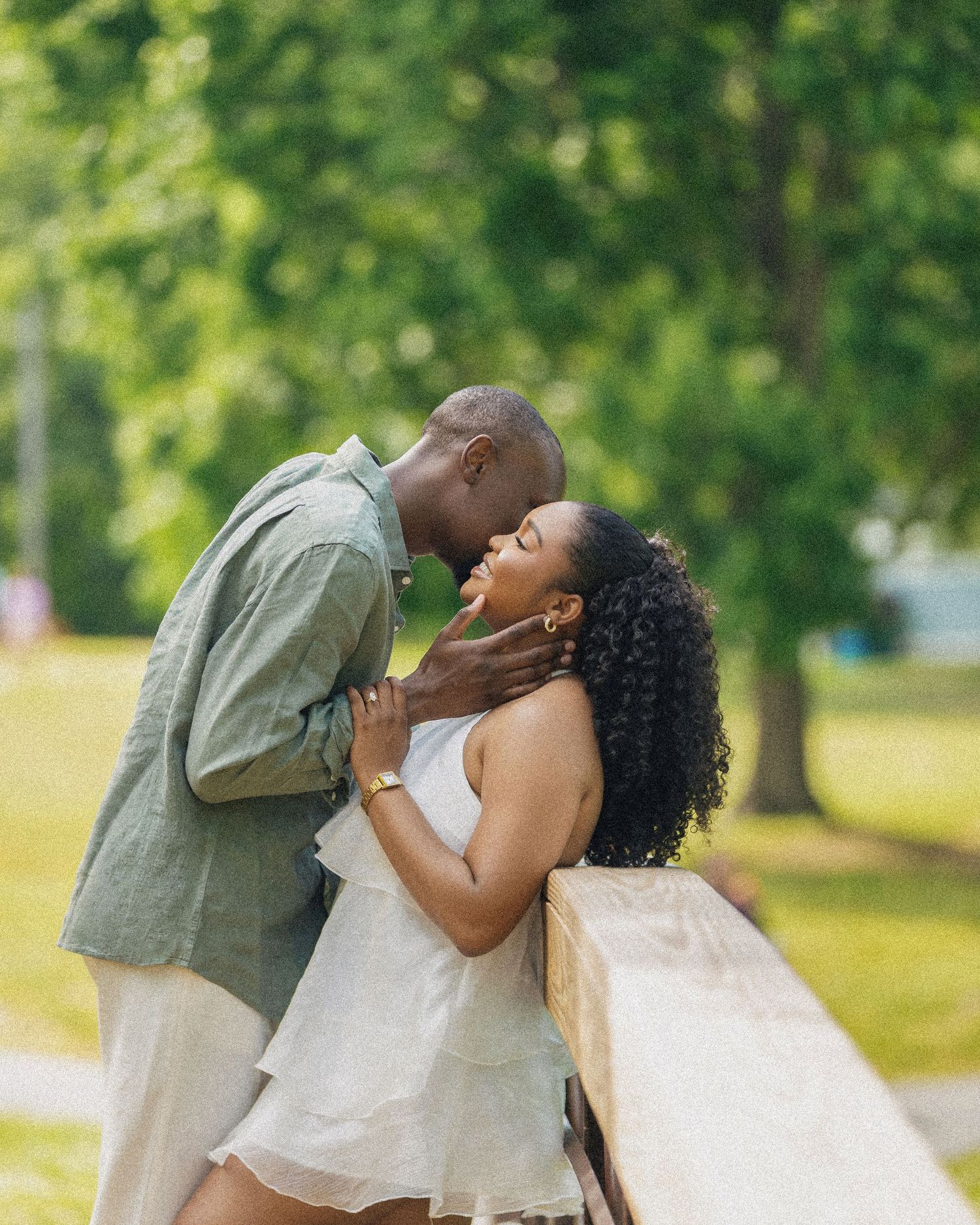 A couple sharing a romantic kiss outdoors in a lush green park, dressed in elegant casual attire.