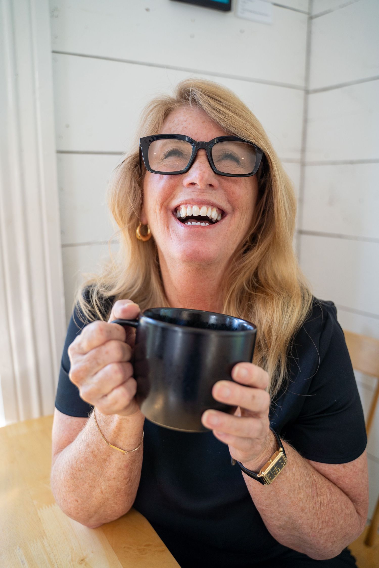 A cheerful middle-aged blonde woman with glasses laughing while holding a black coffee mug at a wooden table.