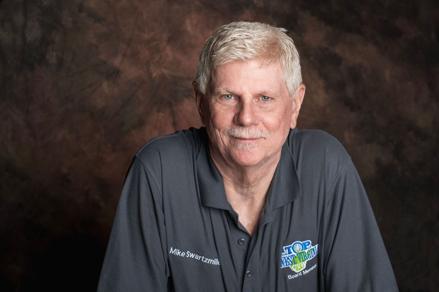Professional headshot of a smiling senior man with gray hair wearing a dark polo shirt with a logo badge.