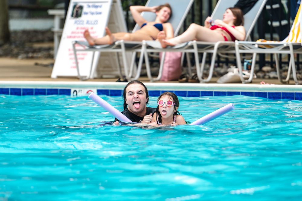 Two people swimming in a pool with foam noodles, smiling and having fun, while others sunbathe on lounge chairs.