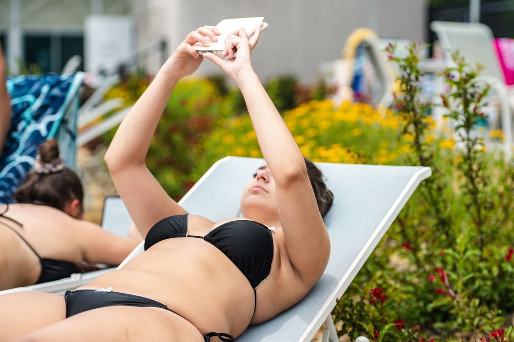 Woman in black bikini lying on lounge chair taking selfie with phone poolside on sunny day.