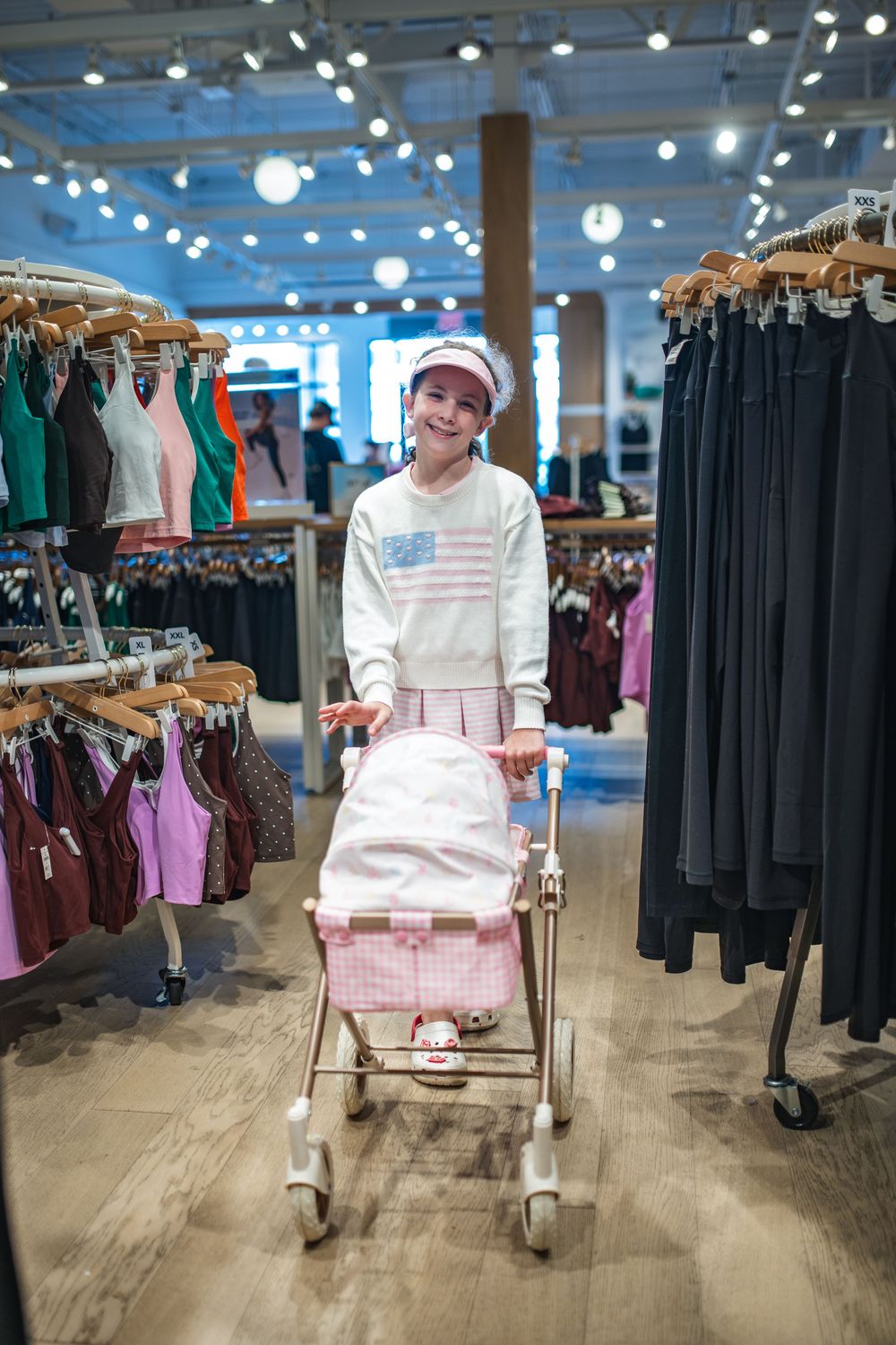 A smiling girl in a white American flag sweatshirt and pink cap pushes a toy baby stroller through a clothing store.