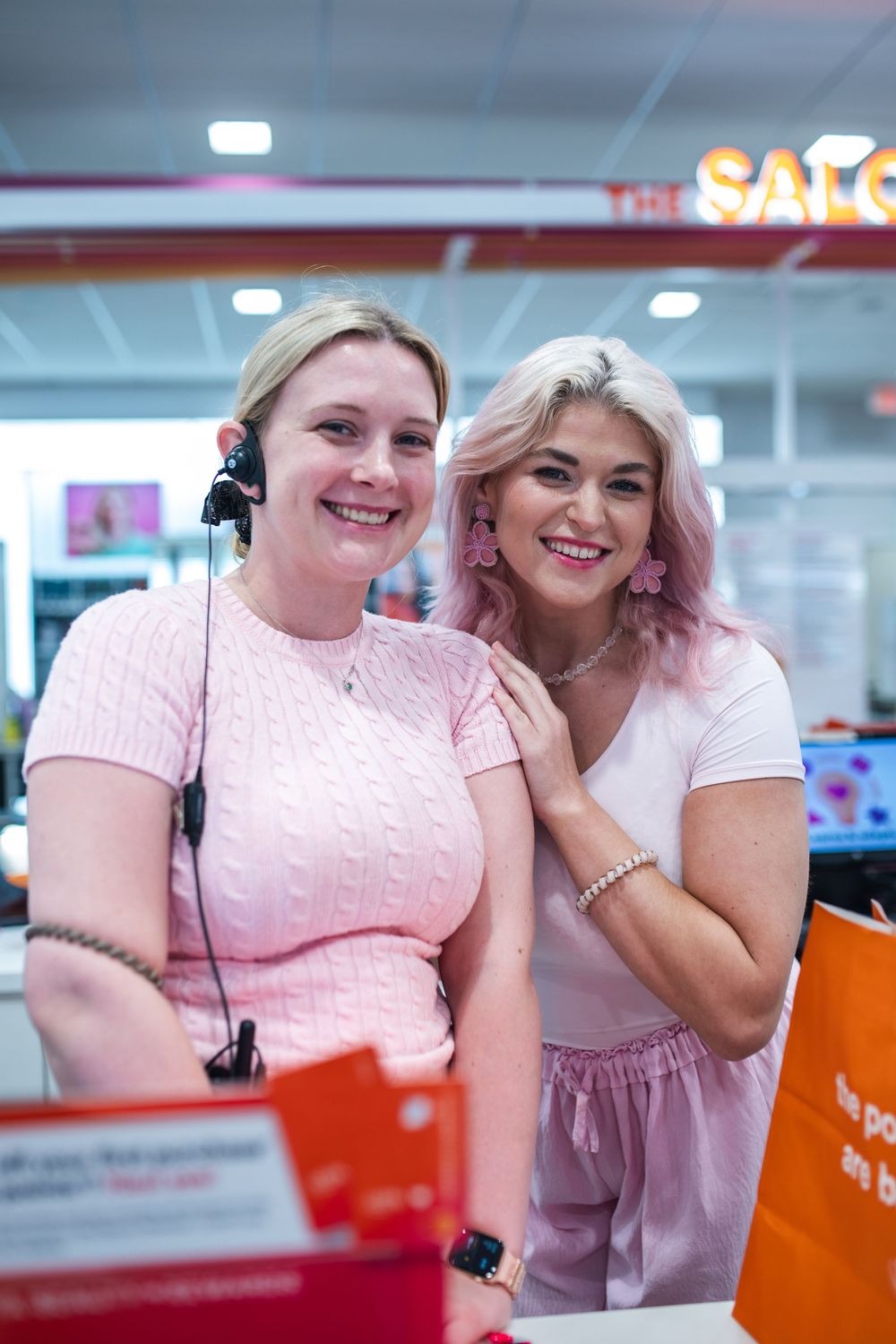 Two smiling women in pink outfits pose together at a retail store counter, one wearing a headset.