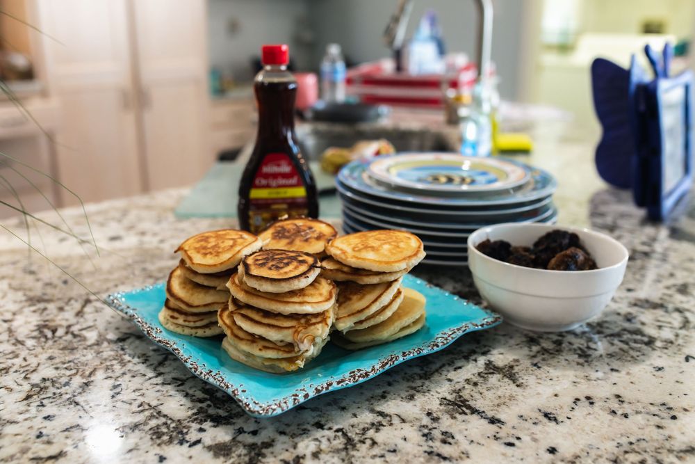 Stack of fluffy pancakes on a blue plate with maple syrup and a bowl of fruit on a granite kitchen counter.
