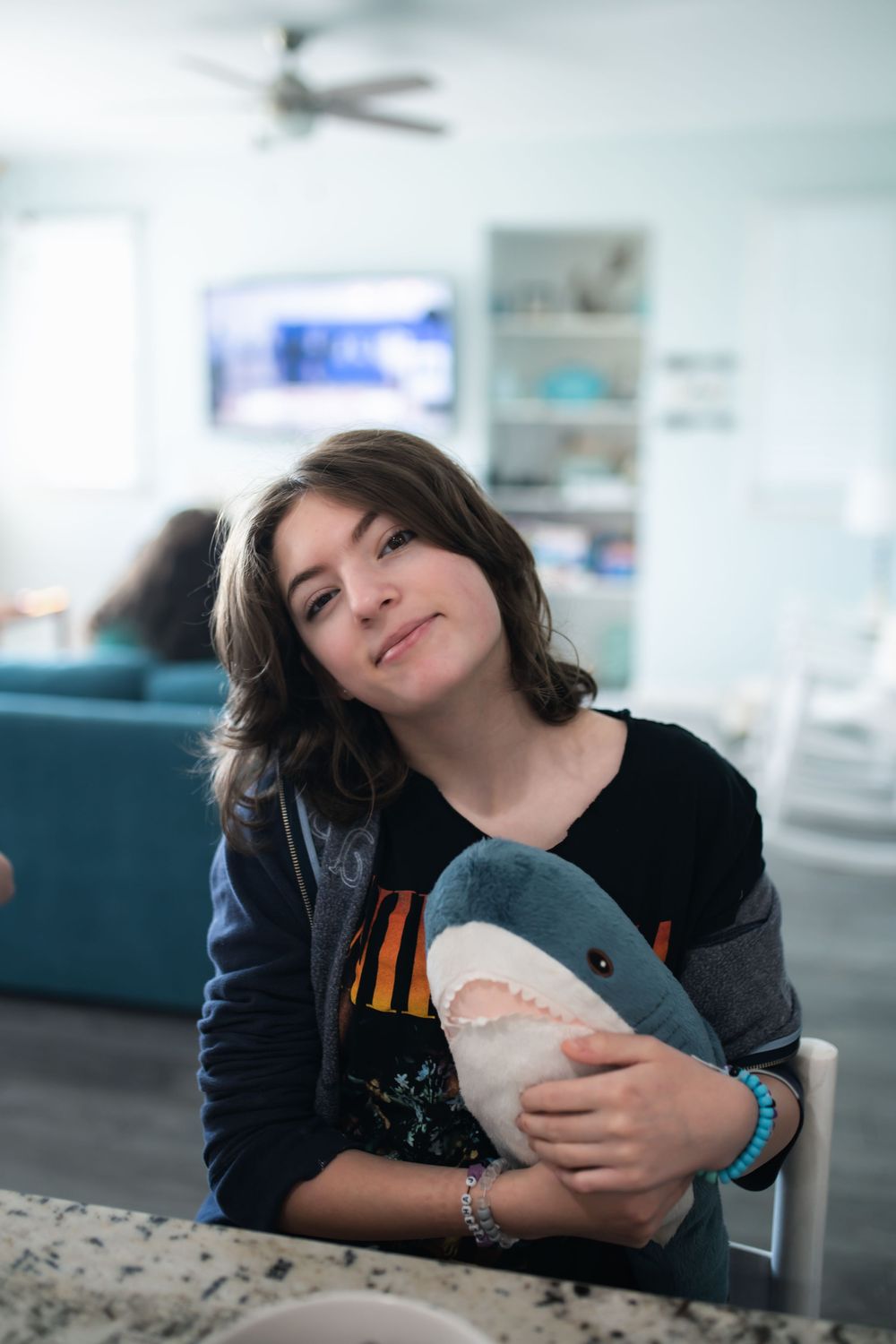 A young teen smiles while hugging a plush shark toy at a kitchen counter in a cozy living space.