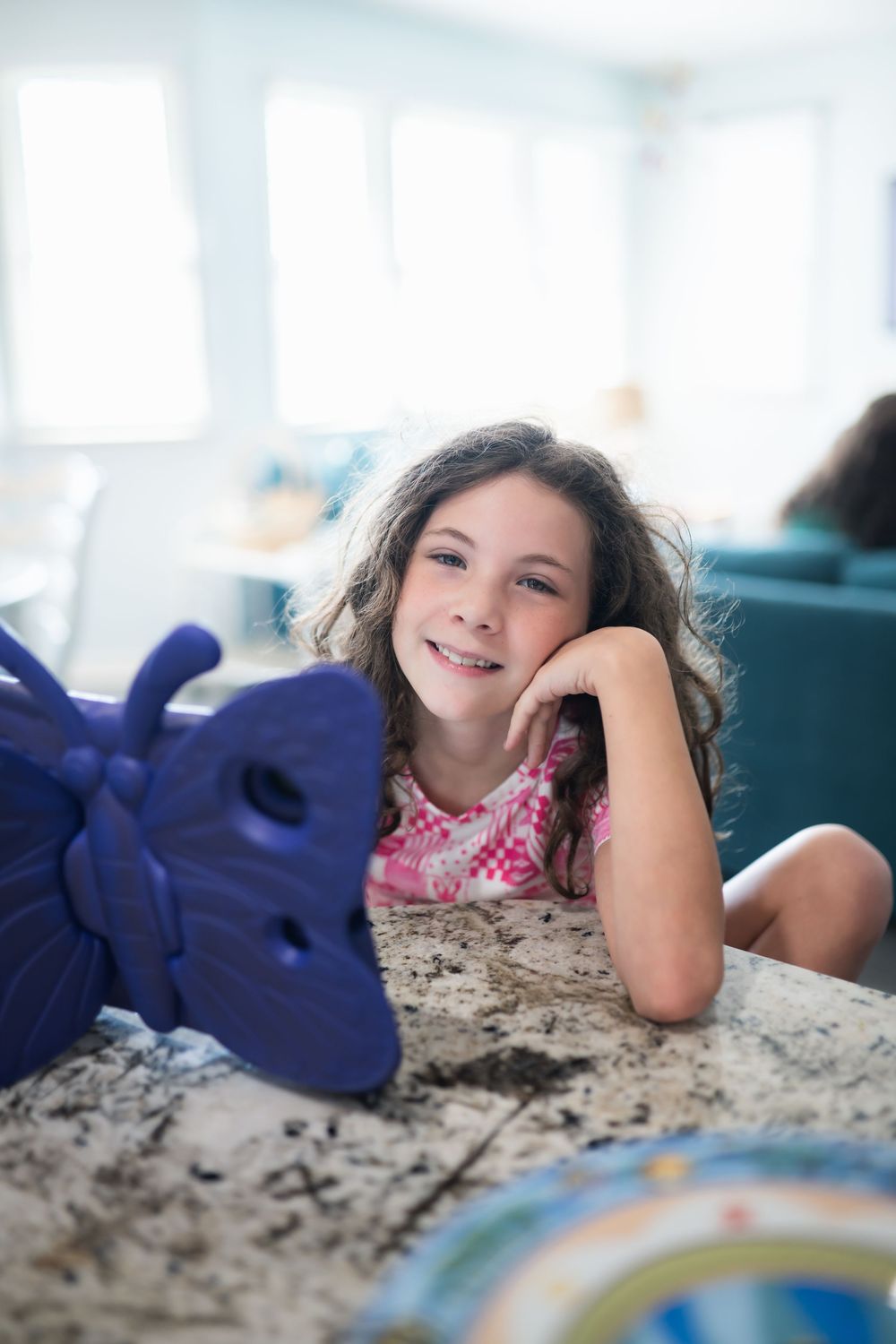 A smiling young girl rests her chin on her hand while lying on a granite countertop next to a blue butterfly toy.