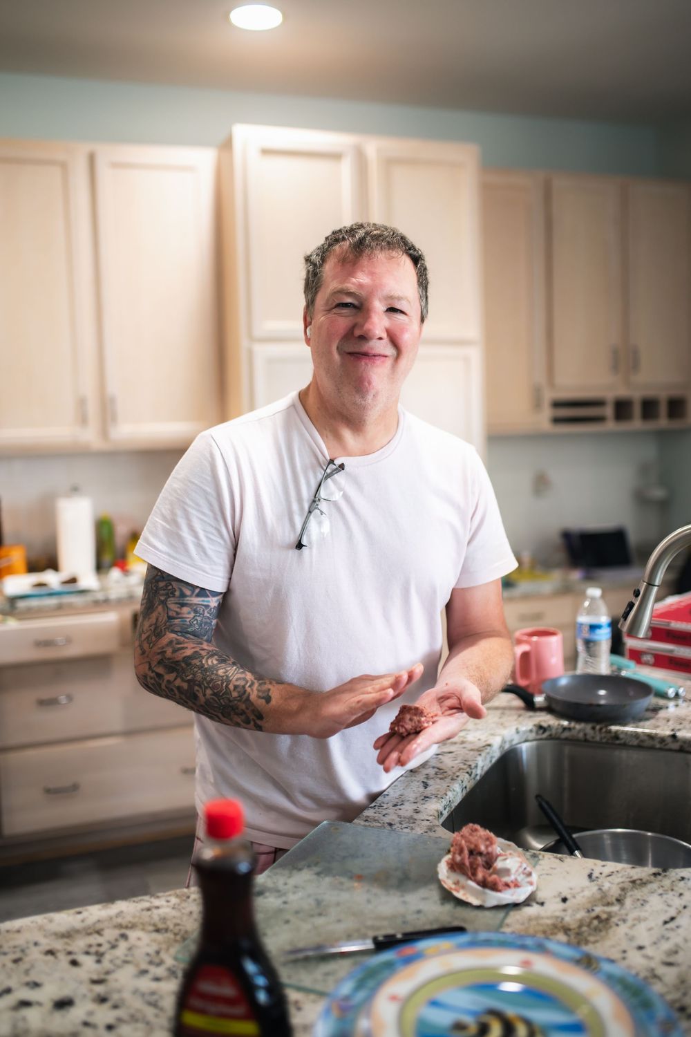 Smiling man with tattoos in white t-shirt preparing meat in a modern kitchen with granite countertops.
