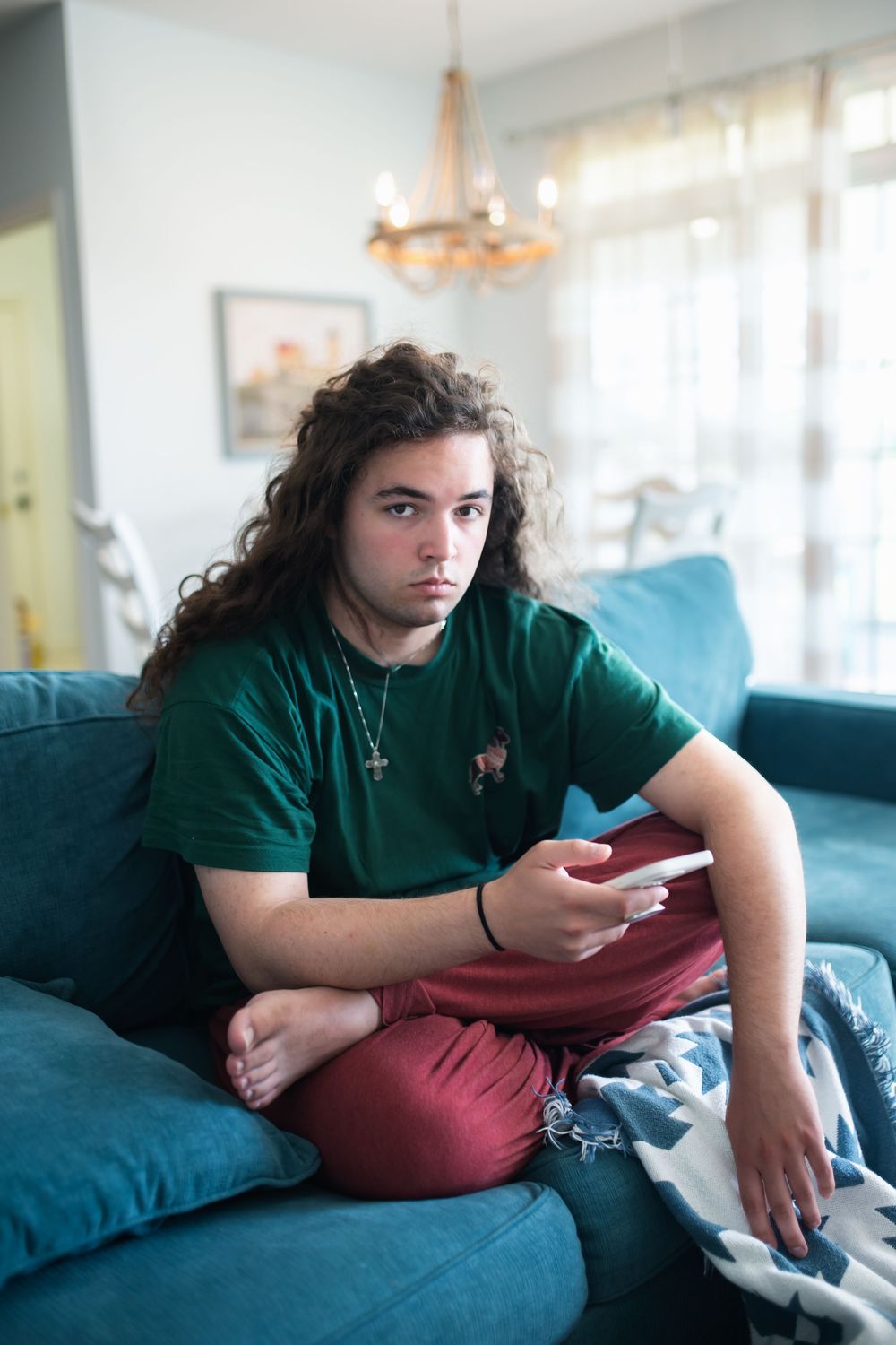 Teen with curly hair sits on blue sofa holding phone, wearing green shirt and red pants indoors.