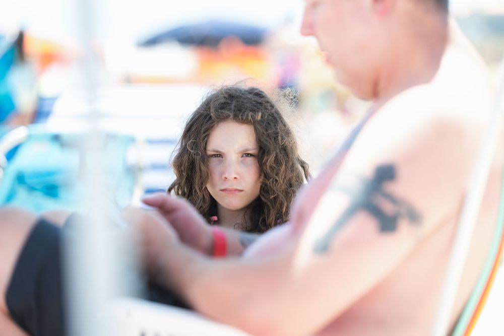 Young girl with curly hair looking seriously at a tattooed man at a sunny beach setting.