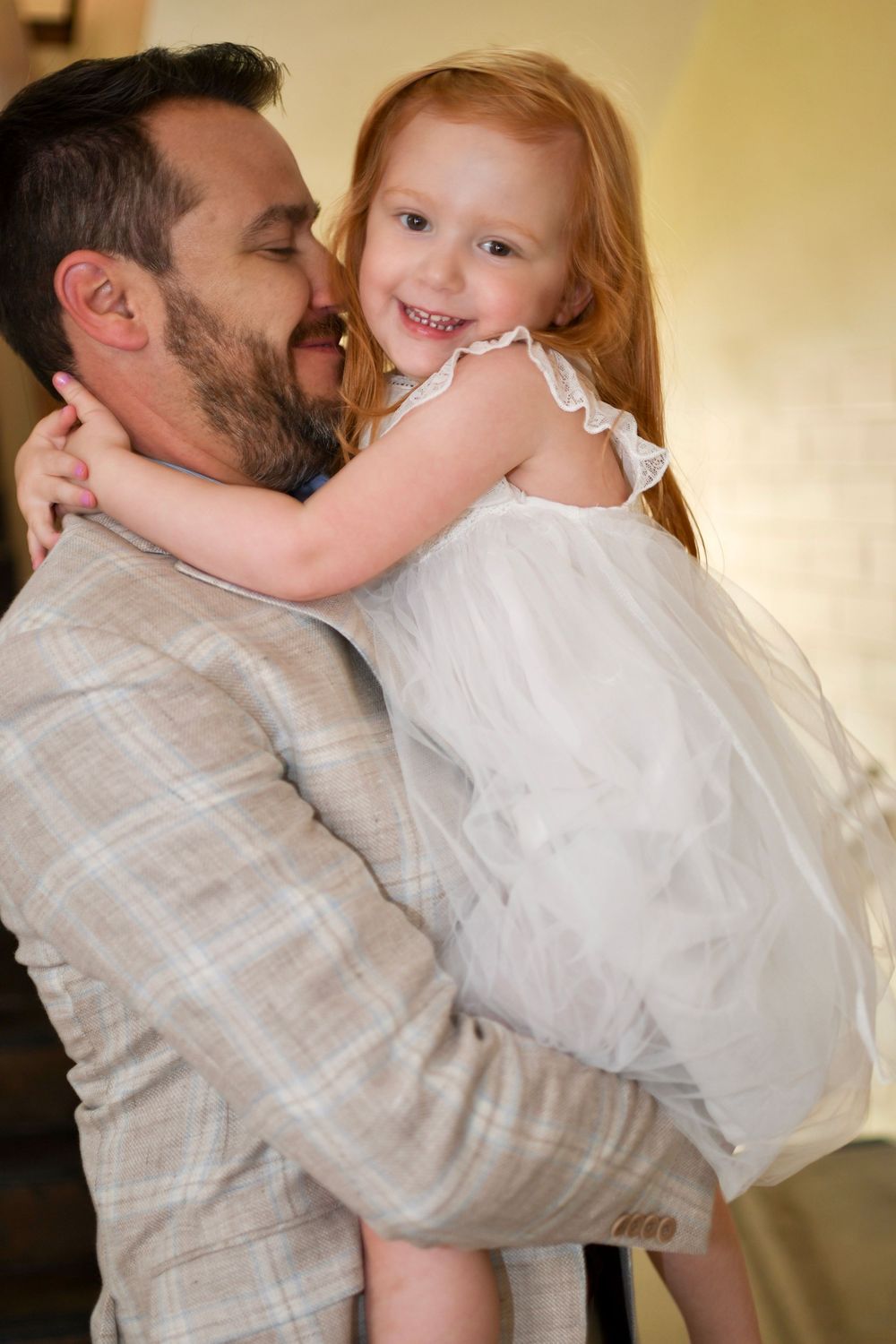 A bearded man in a plaid suit holds a smiling red-haired girl in a white dress, sharing a joyful embrace.