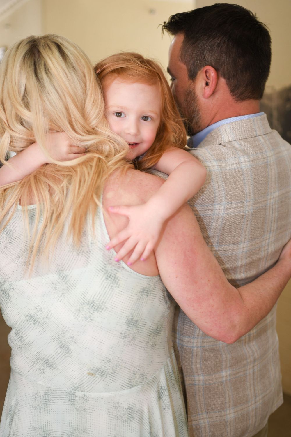 A young redhead girl smiles while being hugged by her blonde mother and dark-haired father indoors.