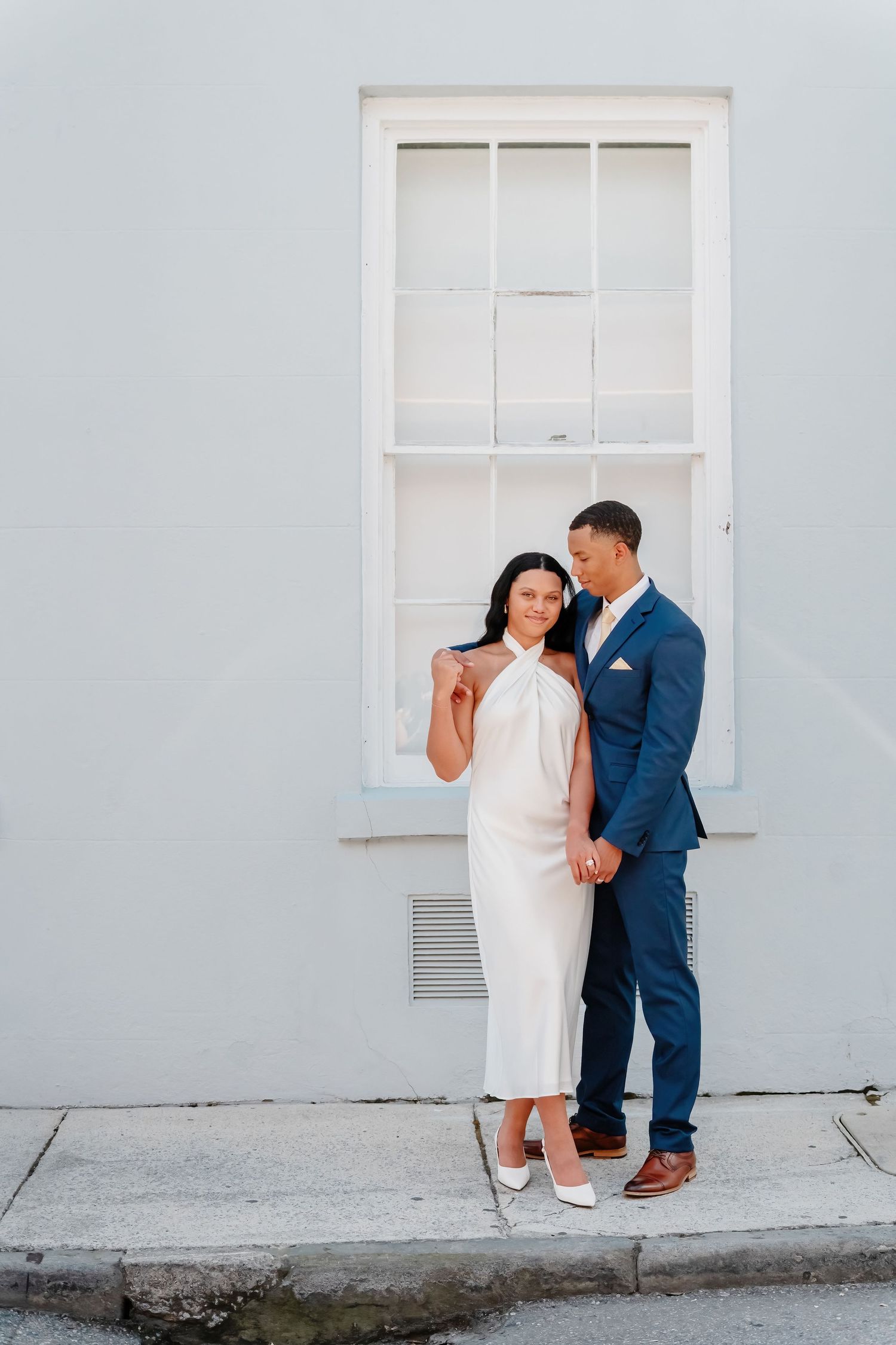 Couple in wedding attire posing in front of a white building, woman in white dress, man in navy suit.