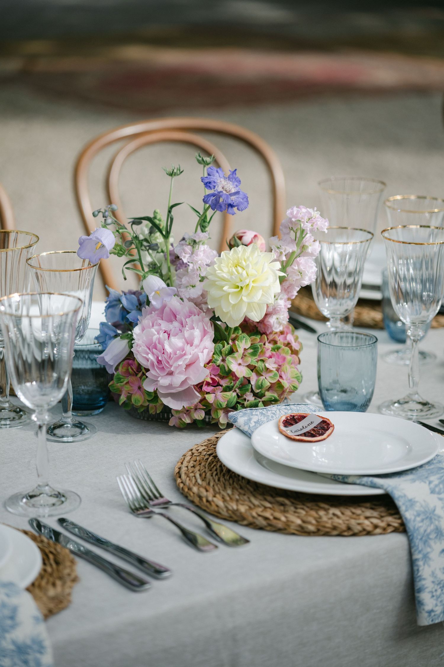 Elegant wedding table setting with pastel floral centerpiece, crystal glasses, woven placemats, and fine china on a gray linen tablecloth.