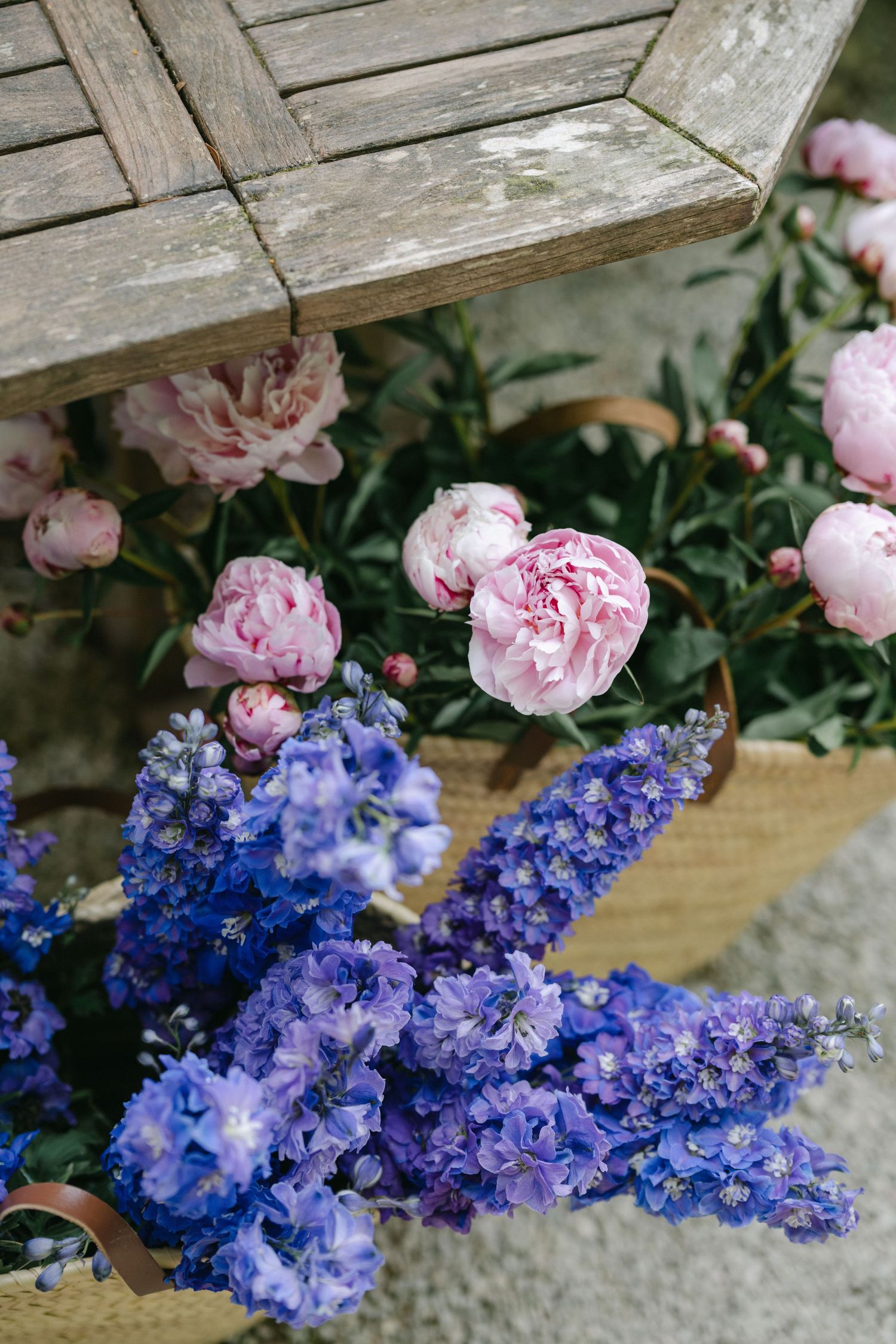Blue delphiniums and pink peonies arranged in a wicker basket near a rustic wooden bench outdoors.